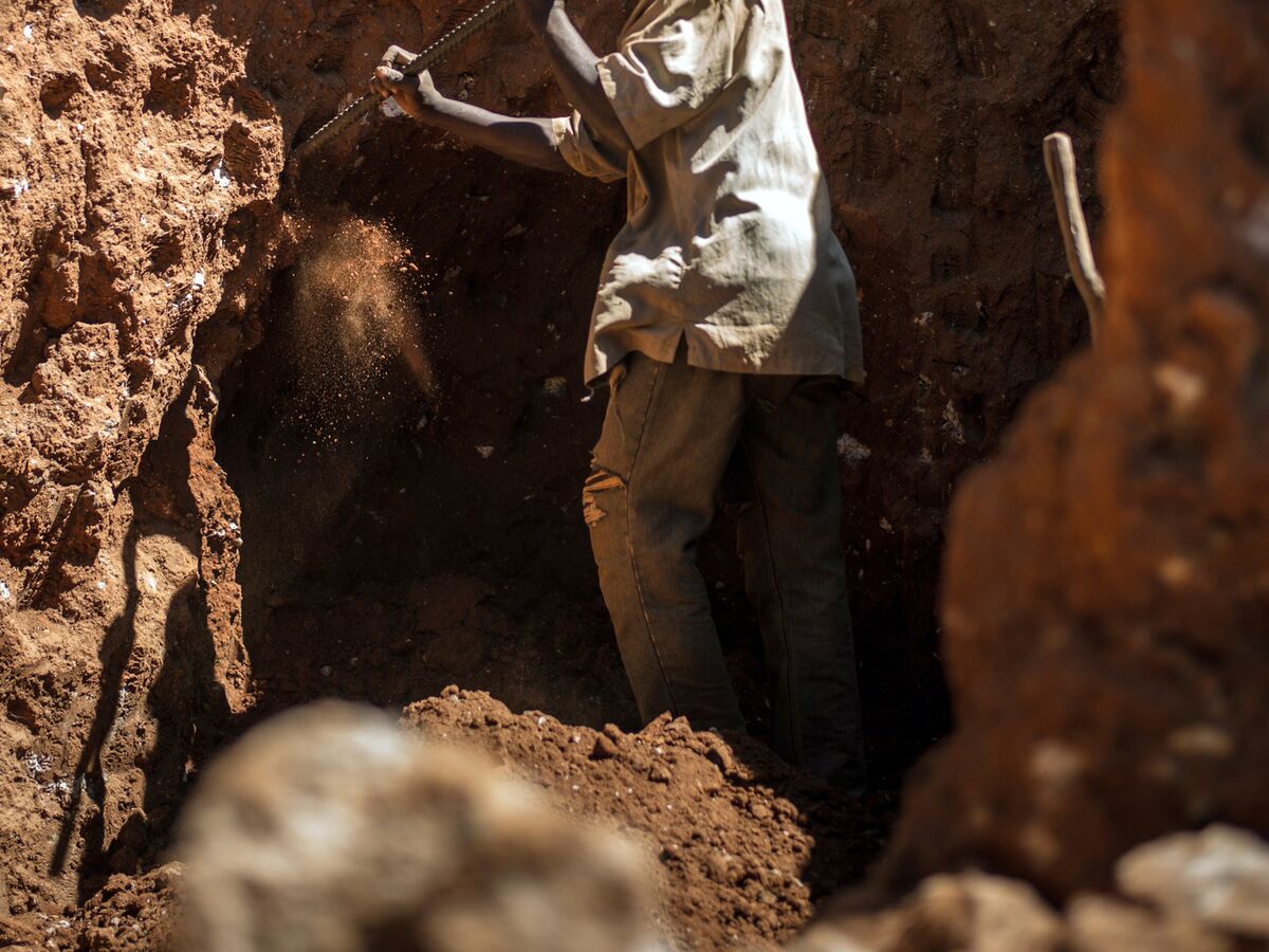 man wearing a black jacket, white shirt, blue jeans, and brown boots, playing a white electric guitar while sitting on an amp