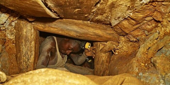 man wearing a black jacket, white shirt, blue jeans, and brown boots, playing a white electric guitar while sitting on an amp