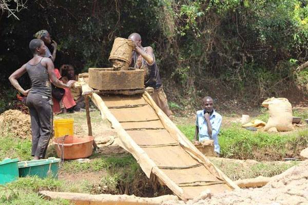 person writing in a notebook beside by an iPad, laptop, printed photos, spectacles, and a cup of coffee on a saucer